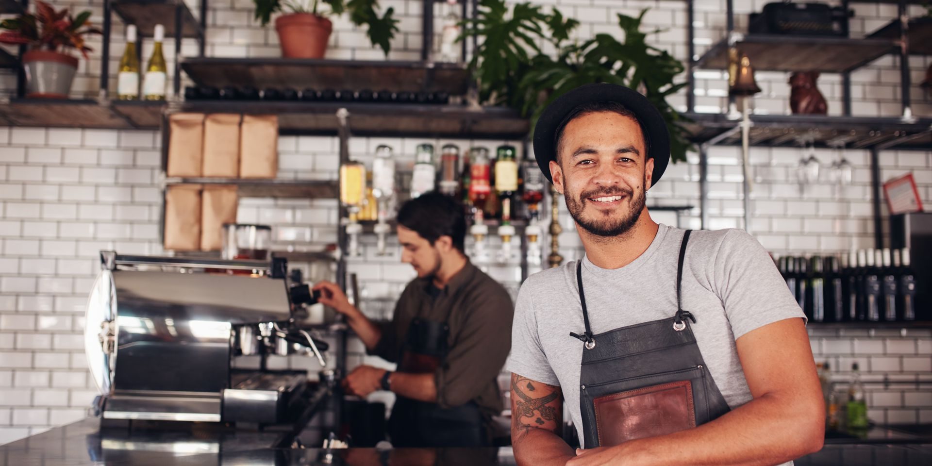 Portrait of male coffee shop owner standing at the counter with barista working in background making drinks.